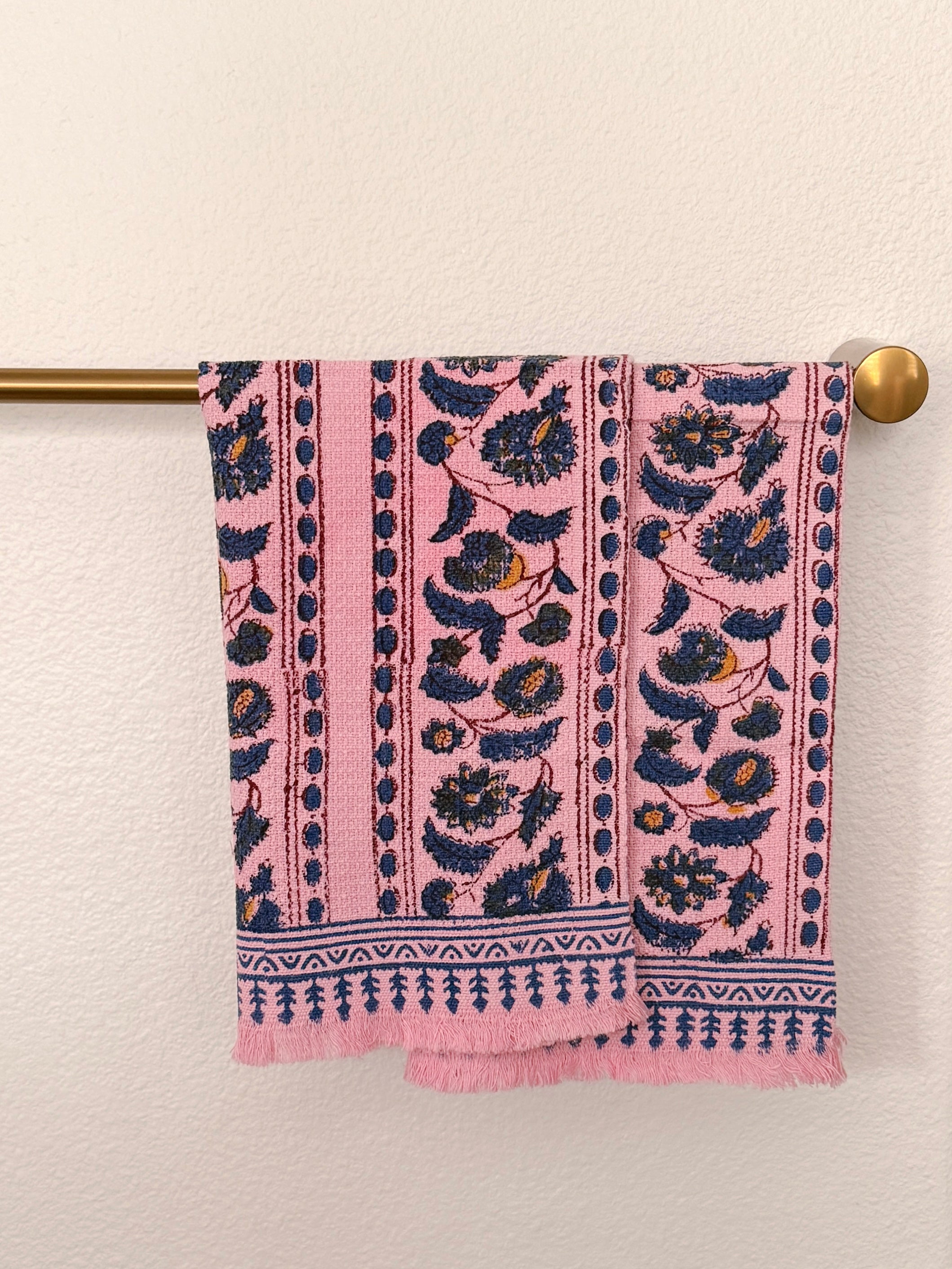 Two pink woven towels with navy blue and mustard yellow floral patterns hangs from a brass rod against a textured off-white wall.