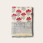 A folded woven bath towel with a cream base, featuring red floral motifs, gray leafy stems, a decorative gray border, and gray diagonal stripes with cream fringe, shown on a light beige background.