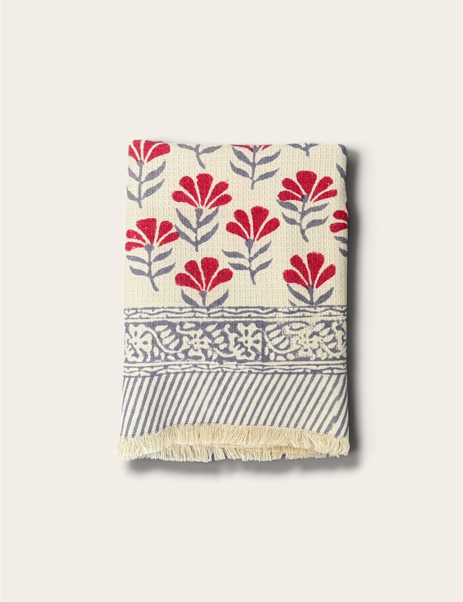 A folded woven bath towel with a cream base, featuring red floral motifs, gray leafy stems, a decorative gray border, and gray diagonal stripes with cream fringe, shown on a light beige background.