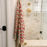 A beige bath towel with a red and gray floral pattern hangs from a brass hook on a white door in a tiled bathroom.
