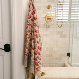 A beige bath towel with a red and gray floral pattern hangs from a brass hook on a white door in a tiled bathroom.