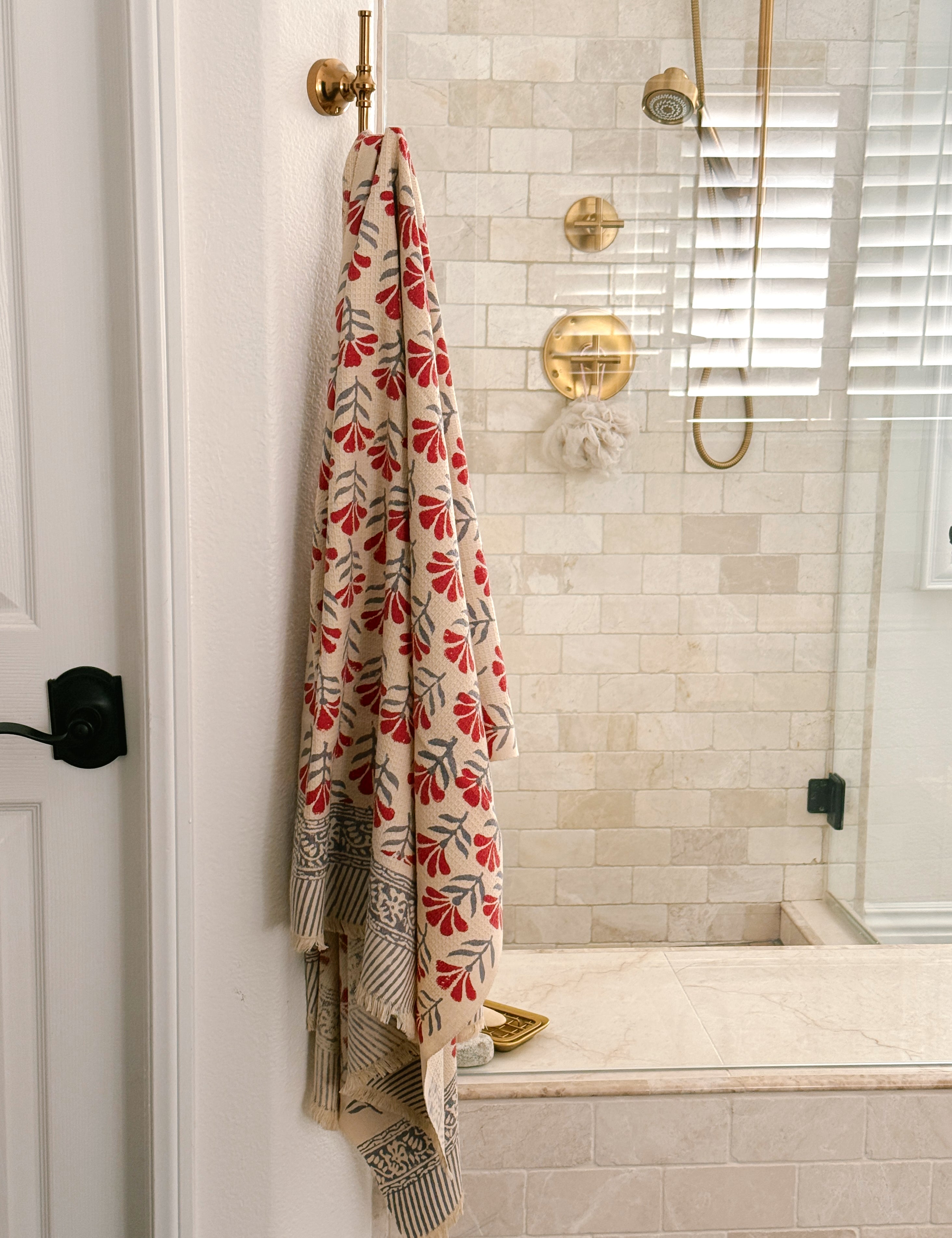 A beige bath towel with a red and gray floral pattern hangs from a brass hook on a white door in a tiled bathroom.