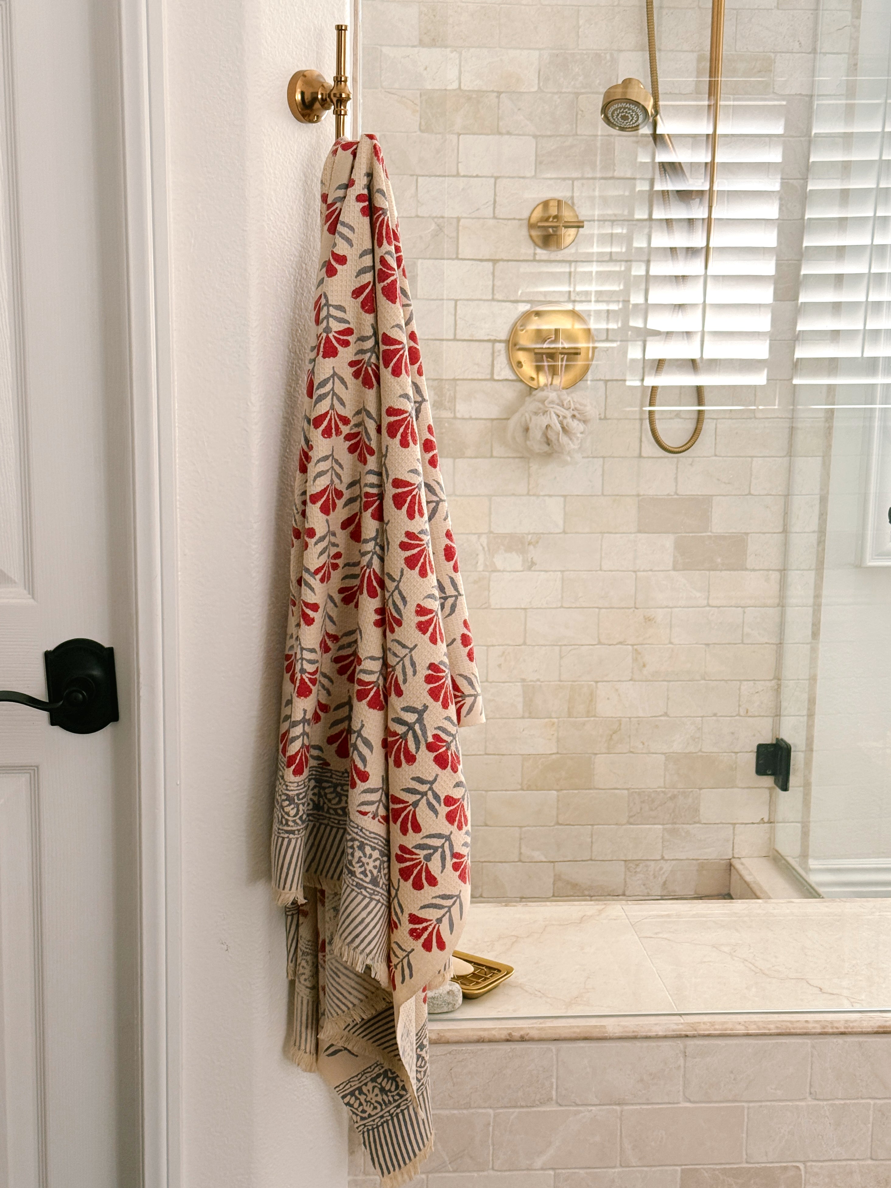A beige bath towel with a red and gray floral pattern hangs from a brass hook on a white door in a tiled bathroom.