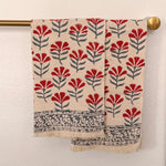 A patterned cotton towel with a red and gray floral block print on a cream background, hanging from a brass rod against a textured white wall.