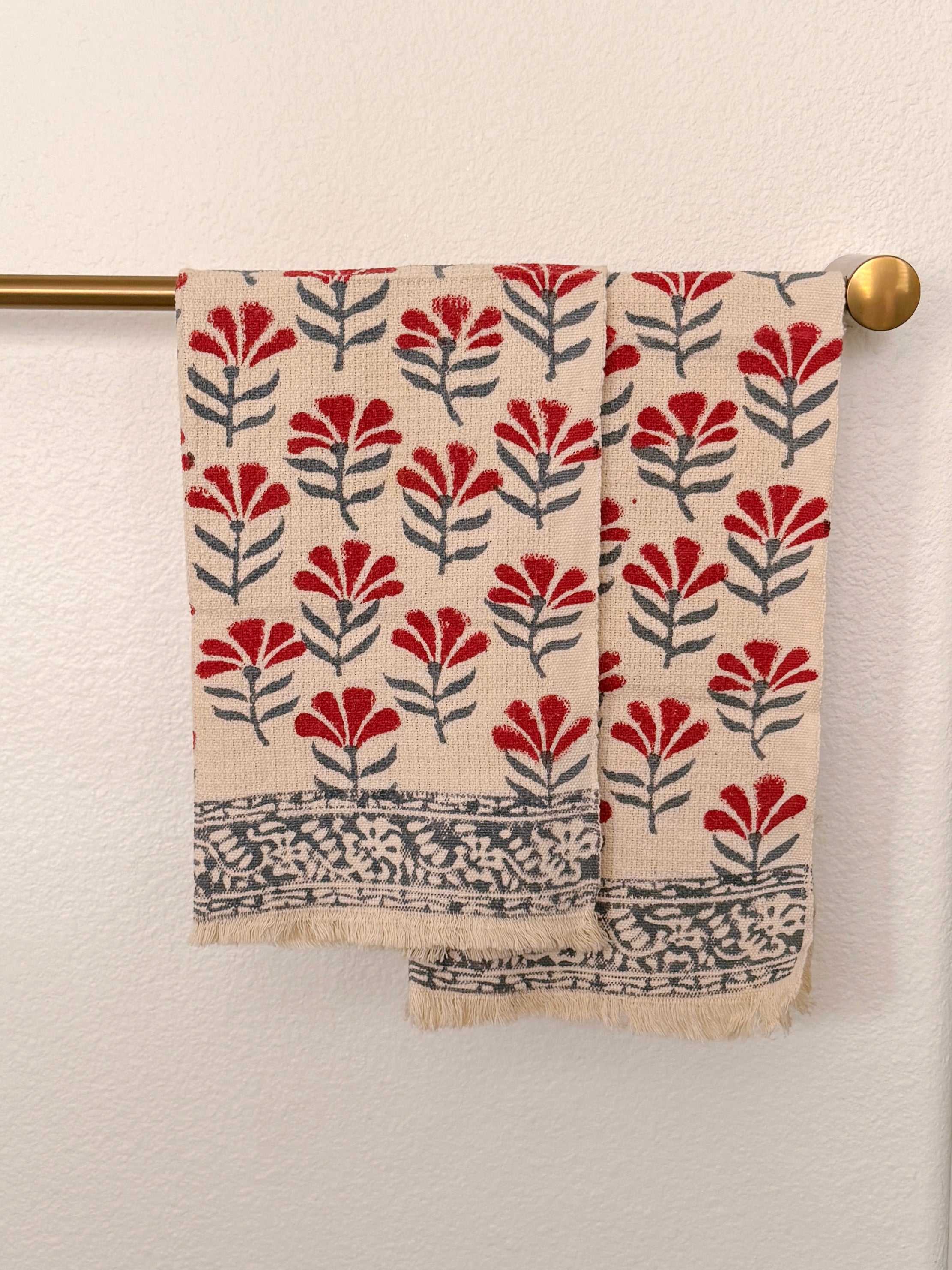 A patterned cotton towel with a red and gray floral block print on a cream background, hanging from a brass rod against a textured white wall.