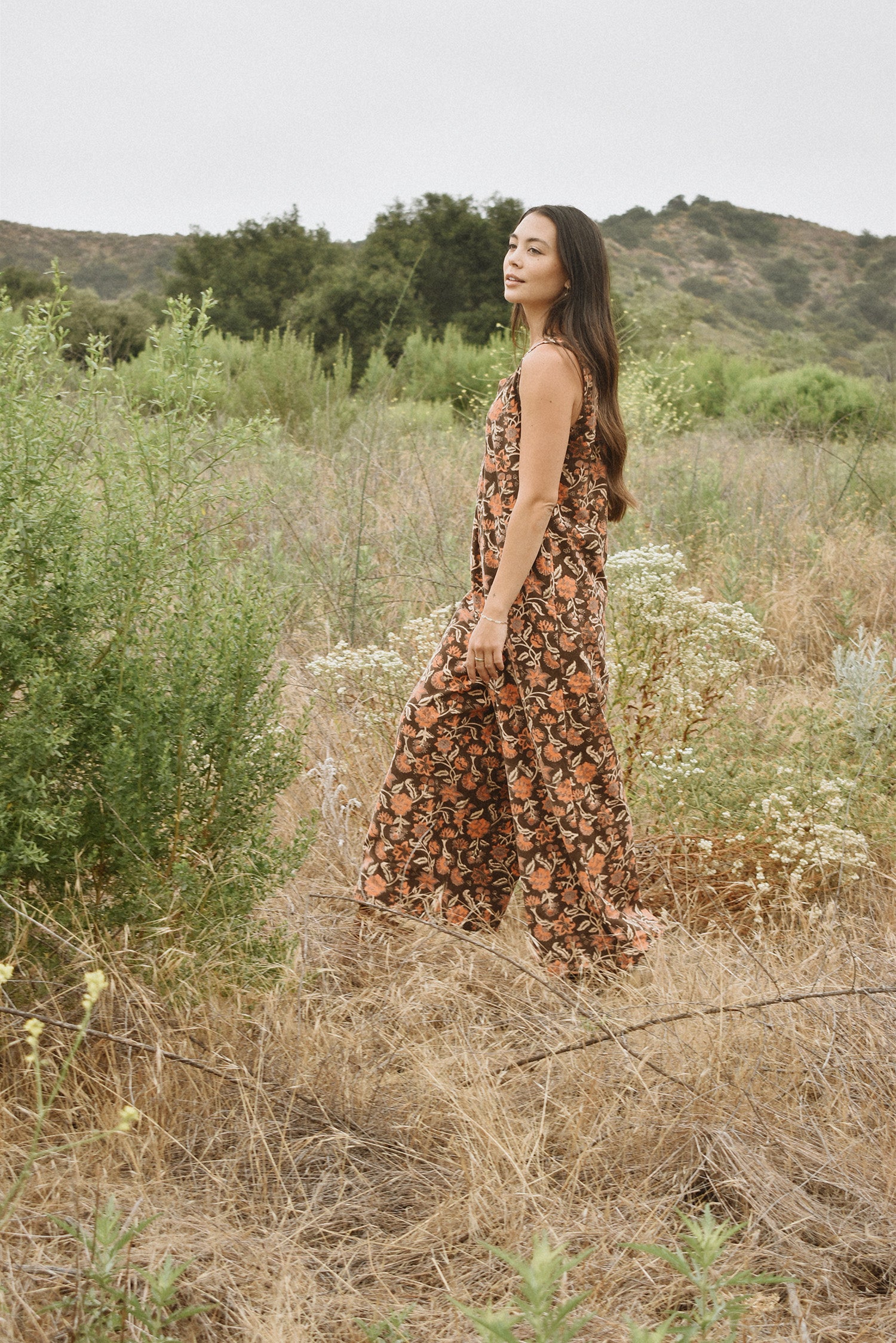 Woman wearing block printed, floral printed Regina corduroy jumpsuit, walking through a field and bushes