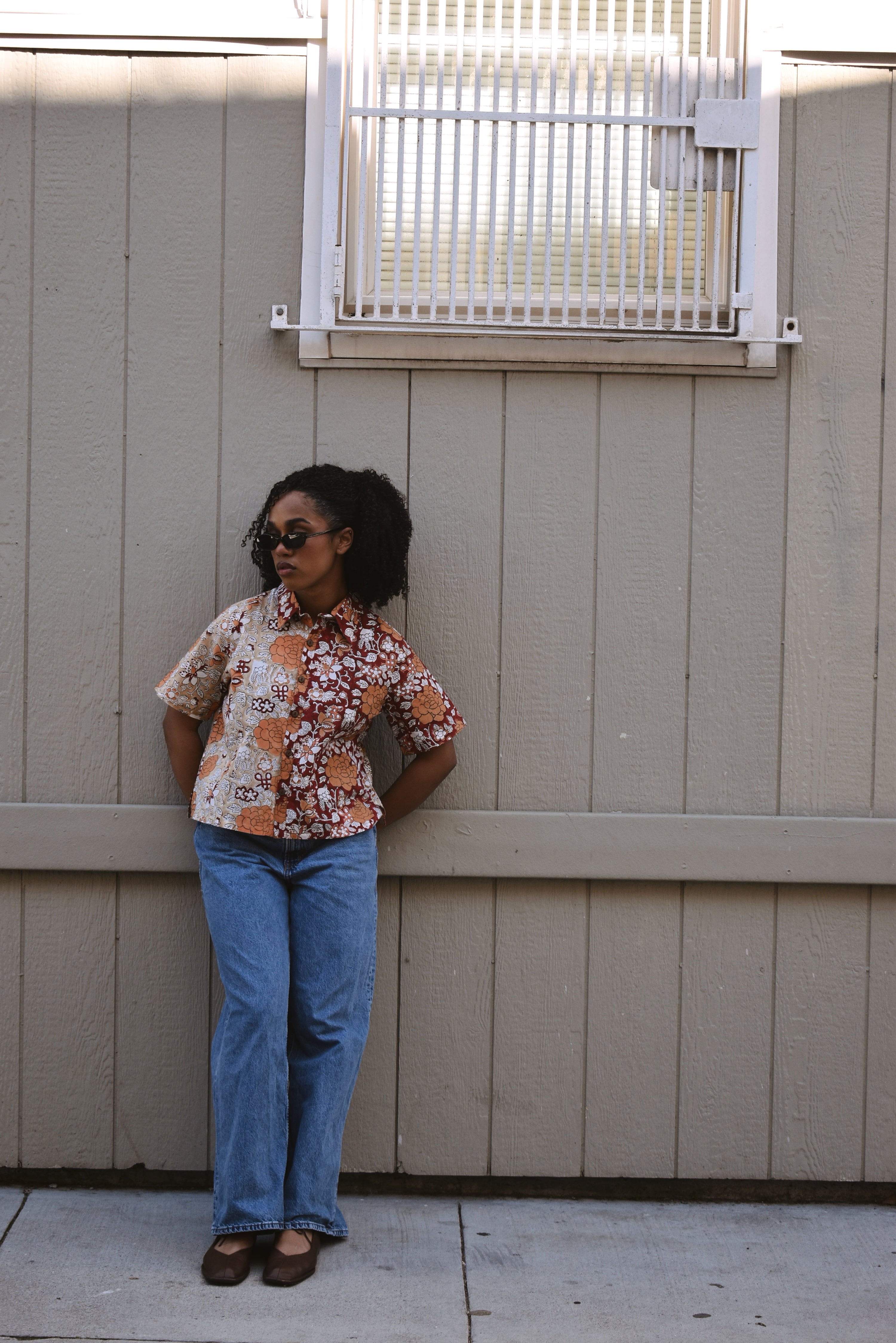 Woman standing outside a building wearing a floral printed shirt and blue jeans with sunglasses