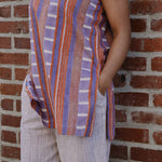 Person wearing a sleeveless red and purple striped top and pant set, standing against a brick wall. Close up on side pant pocket. 