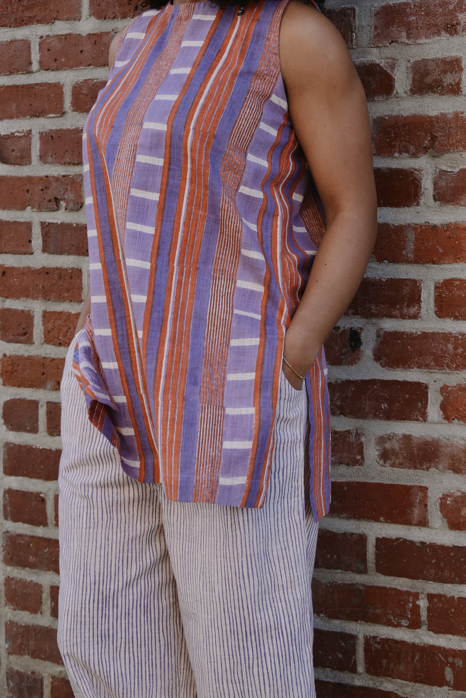 Person wearing a sleeveless red and purple striped top and pant set, standing against a brick wall. Close up on side pant pocket. 