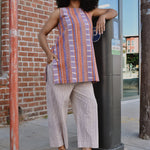 Woman in a striped sleeveless top and pants standing on a sidewalk with arm on a parking meter, in front of a brick wall