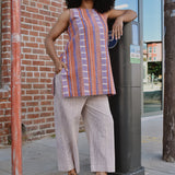 Woman in a striped sleeveless top and pants standing on a sidewalk with arm on a parking meter, in front of a brick wall