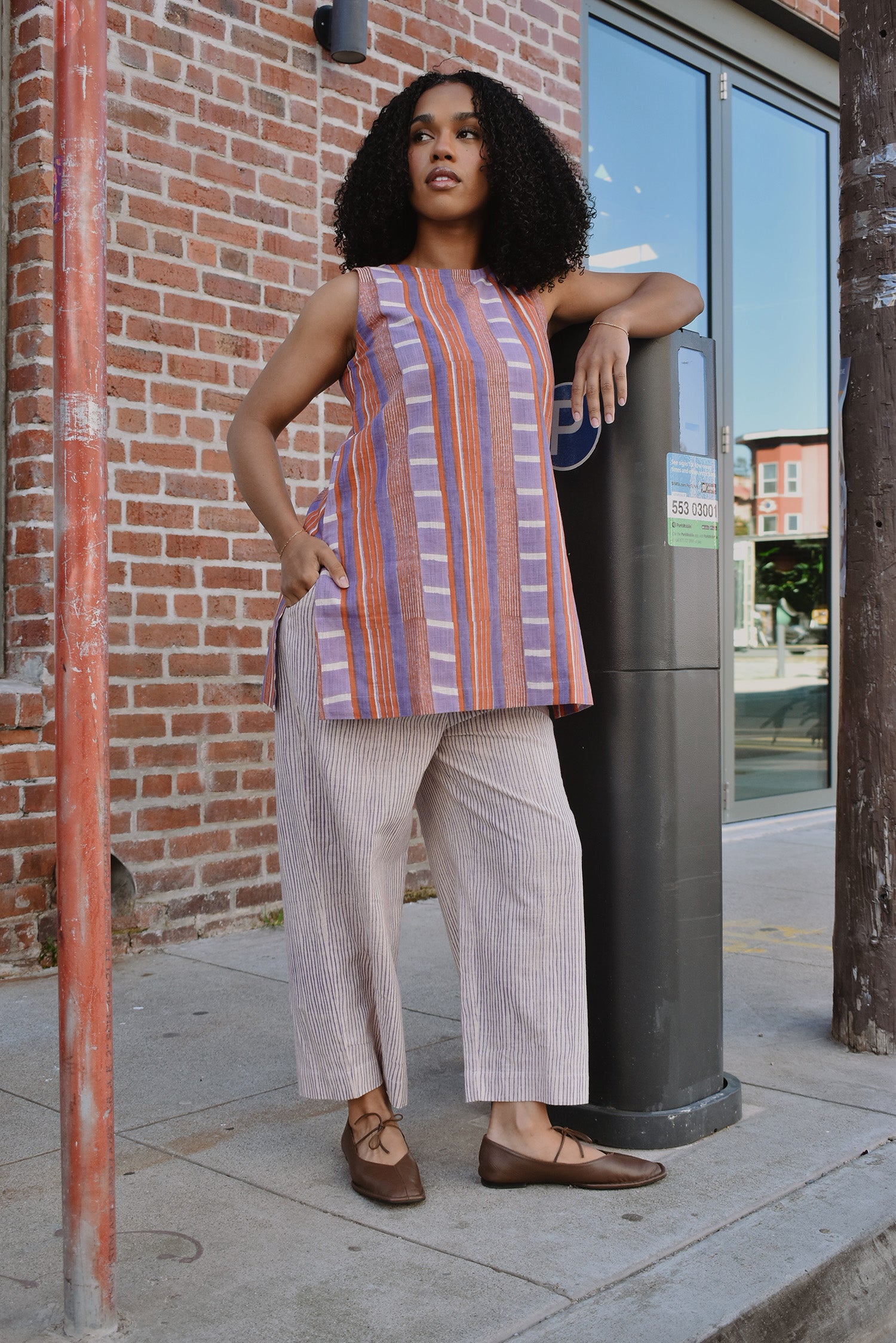 Woman in a striped sleeveless top and pants standing on a sidewalk with arm on a parking meter, in front of a brick wall