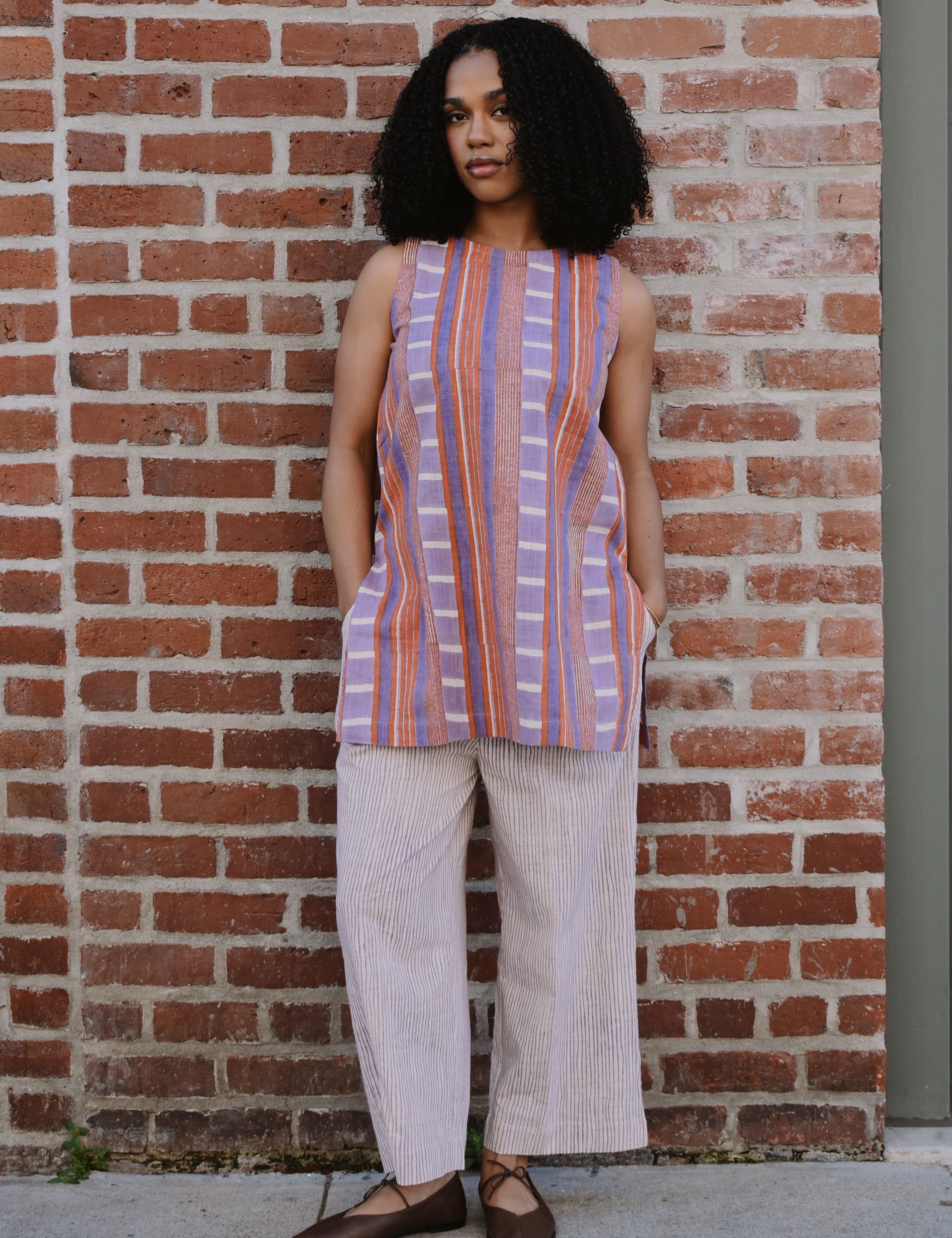 Woman wearing a red and purple striped sleeveless top and wide-leg pants standing against a brick wall.