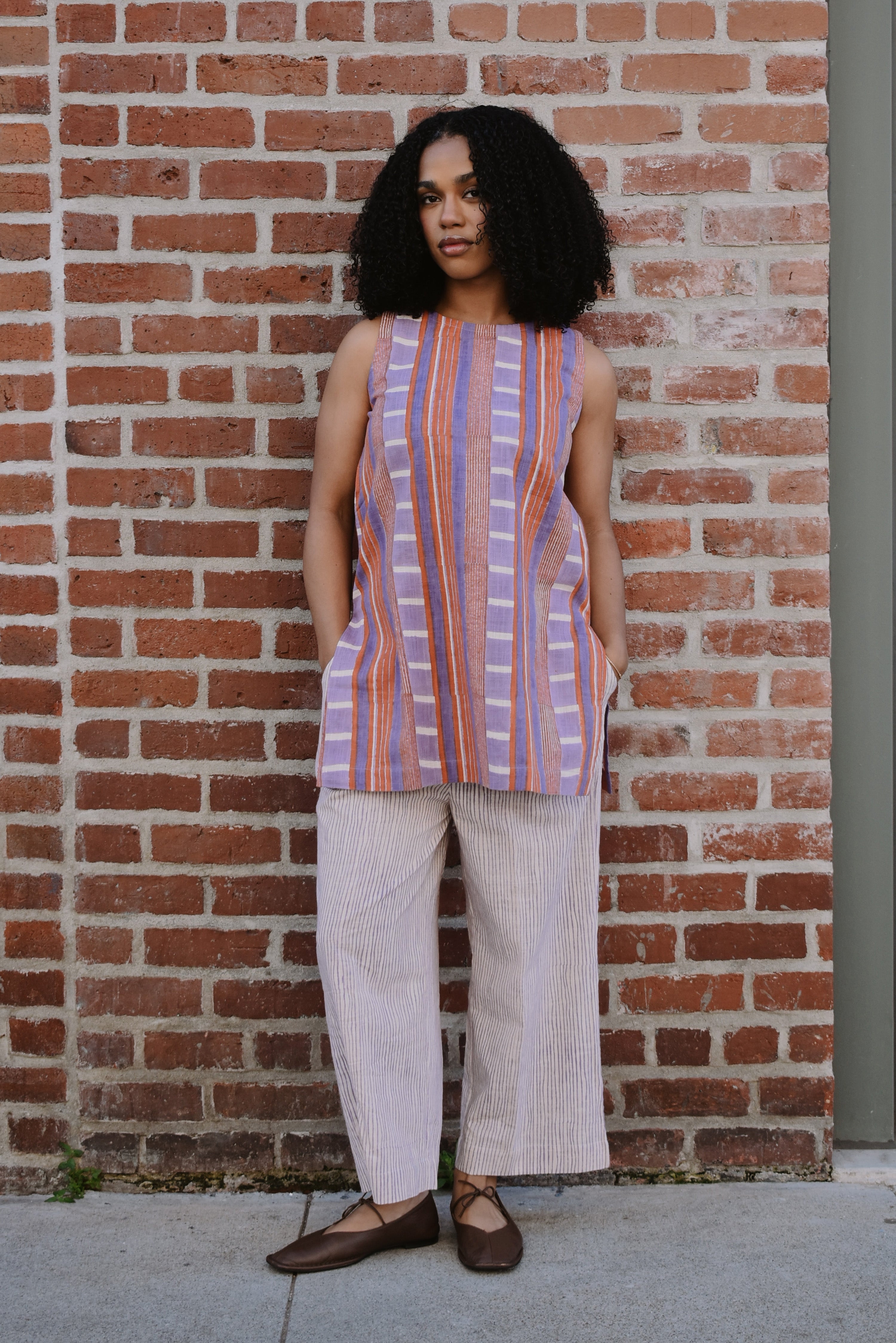 Woman wearing a red and purple striped sleeveless top and wide-leg pants standing against a brick wall.