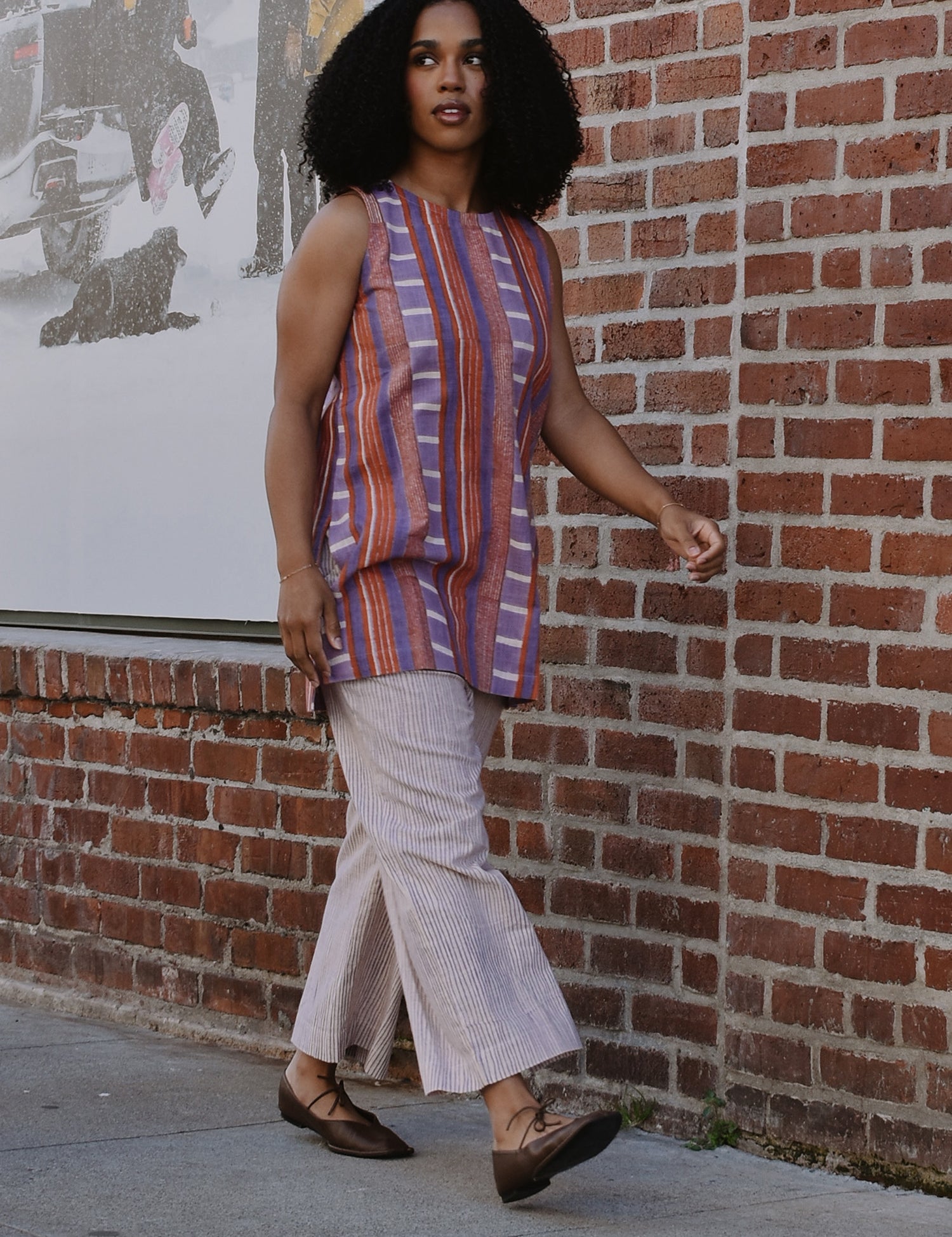 Woman in a red and purple striped sleeveless top and pants walking by a brick wall.