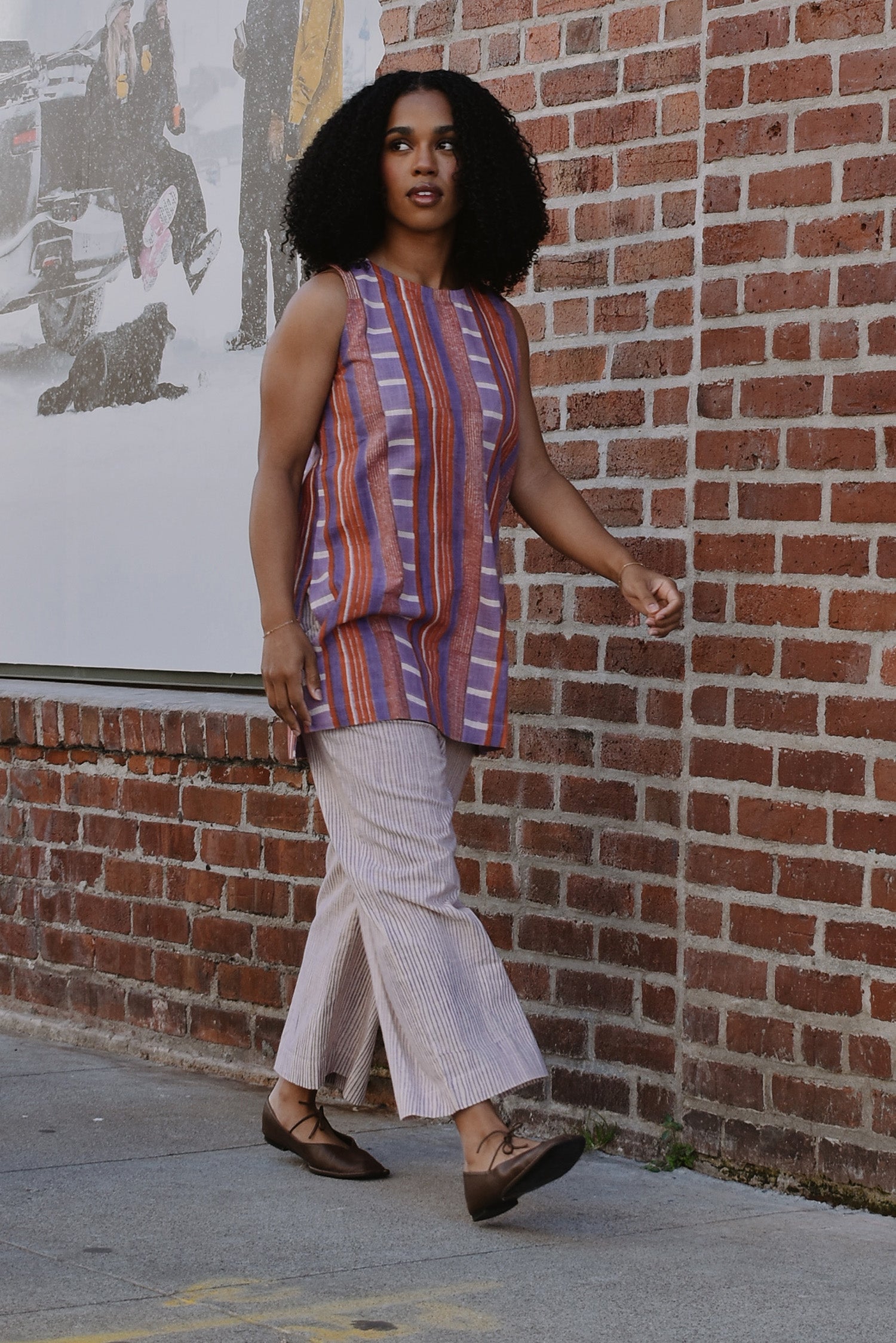 Woman in a red and purple striped sleeveless top and pants walking by a brick wall.