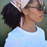 Woman wearing a tan bandana headscarf with a brown floral pattern, sunglasses, and a white shirt against a green background.