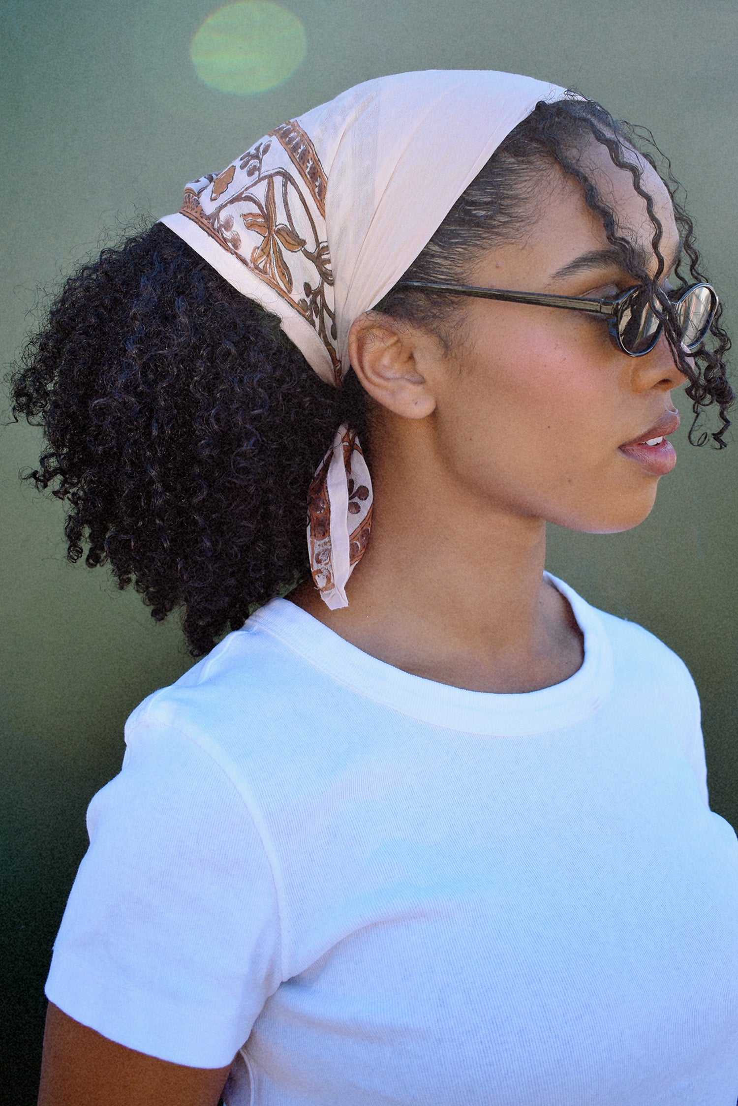 Woman wearing a tan bandana headscarf with a brown floral pattern, sunglasses, and a white shirt against a green background.