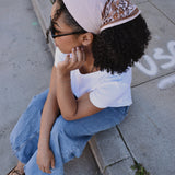 Woman sitting on a sidewalk wearing a floral printed bandana headscarf and white shirt.