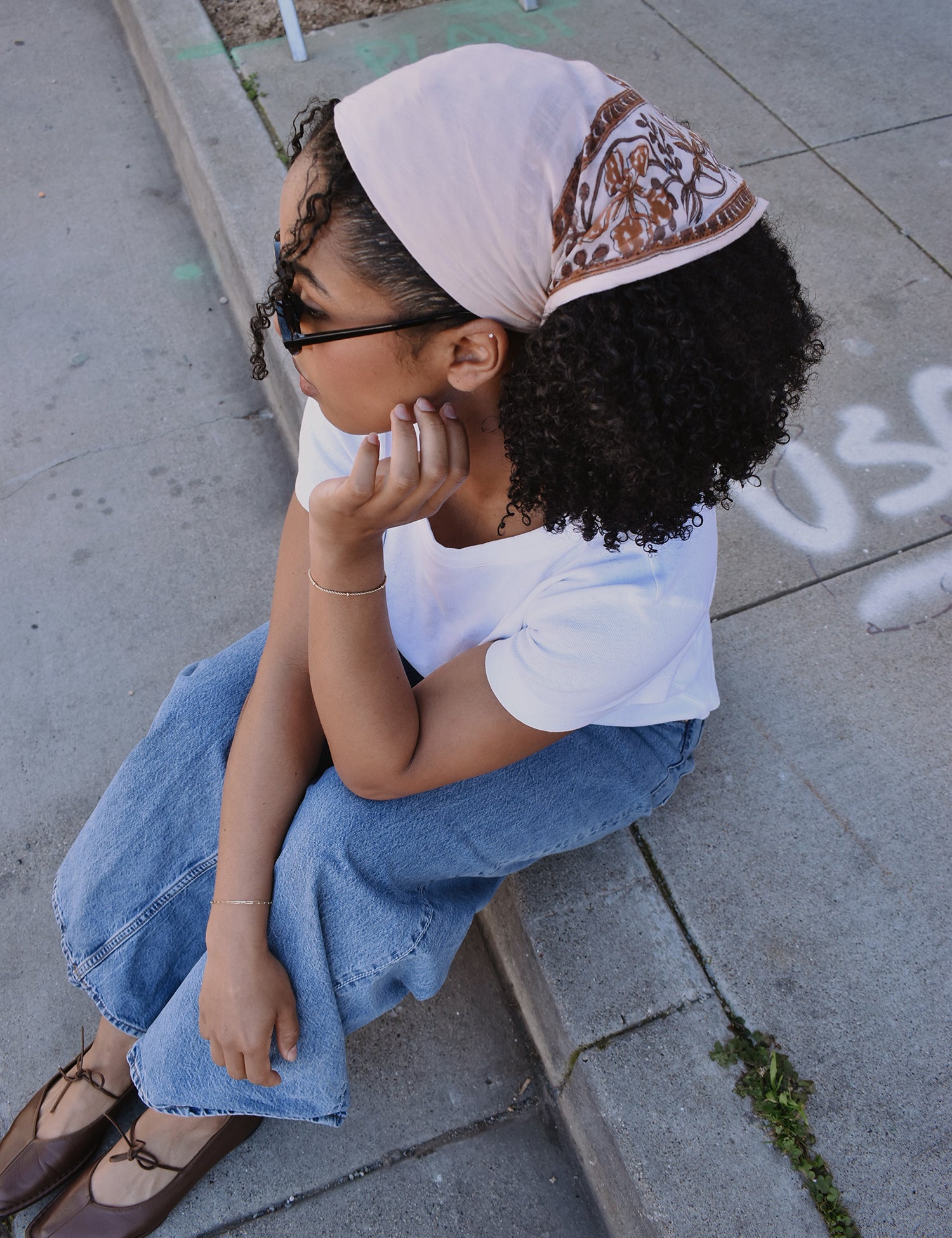 Woman sitting on a sidewalk wearing a floral printed bandana headscarf and white shirt.