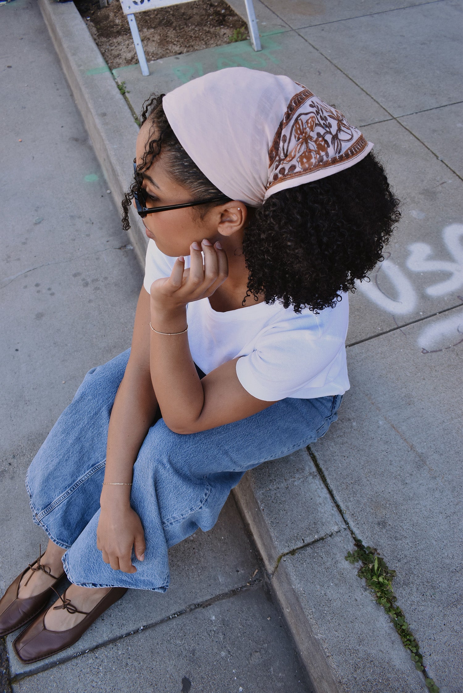 Woman sitting on a sidewalk wearing a floral printed bandana headscarf and white shirt.