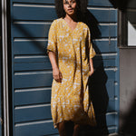 Woman in a yellow floral midi dress standing in front of a blue wall.