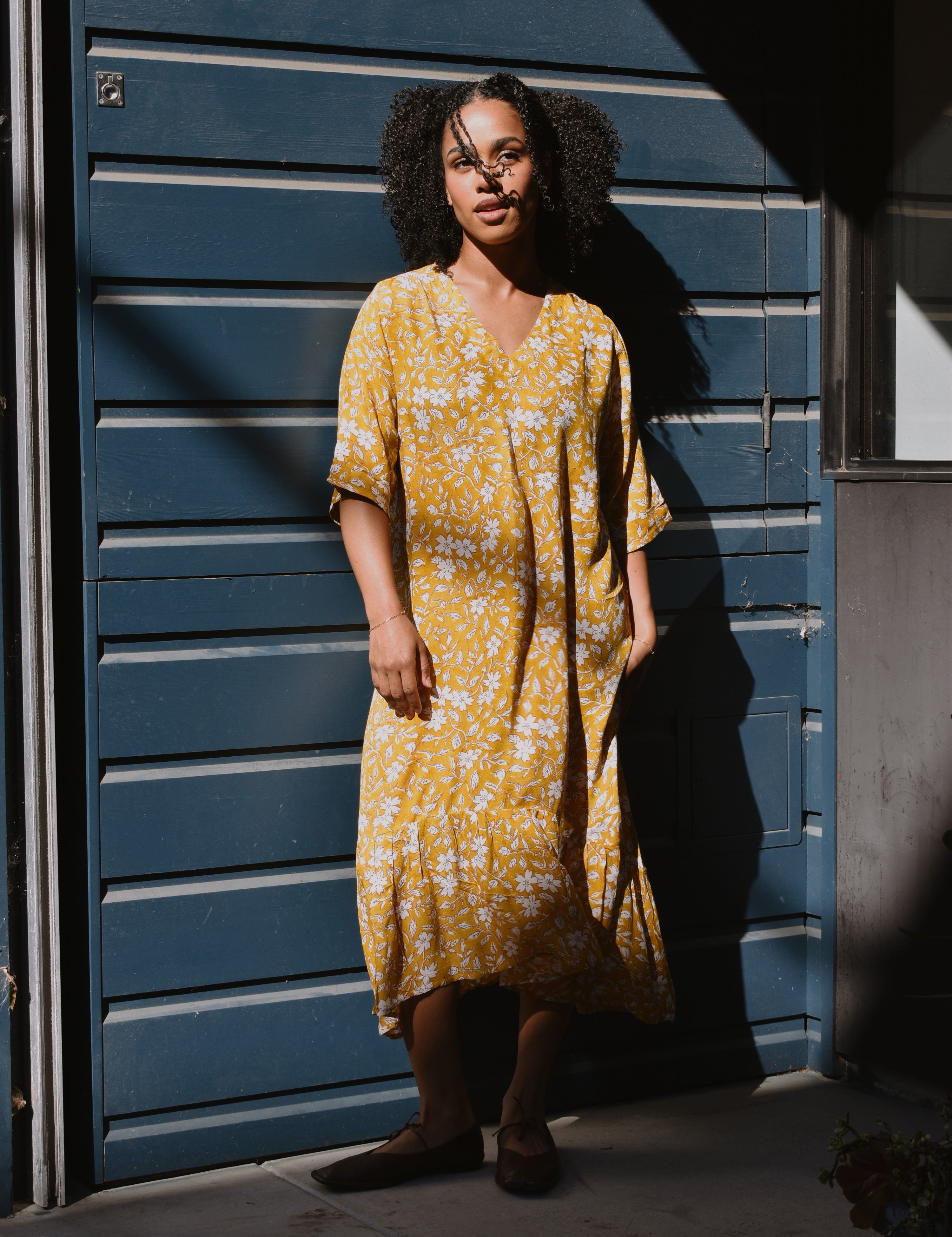 Woman in a yellow floral midi dress standing in front of a blue wall.