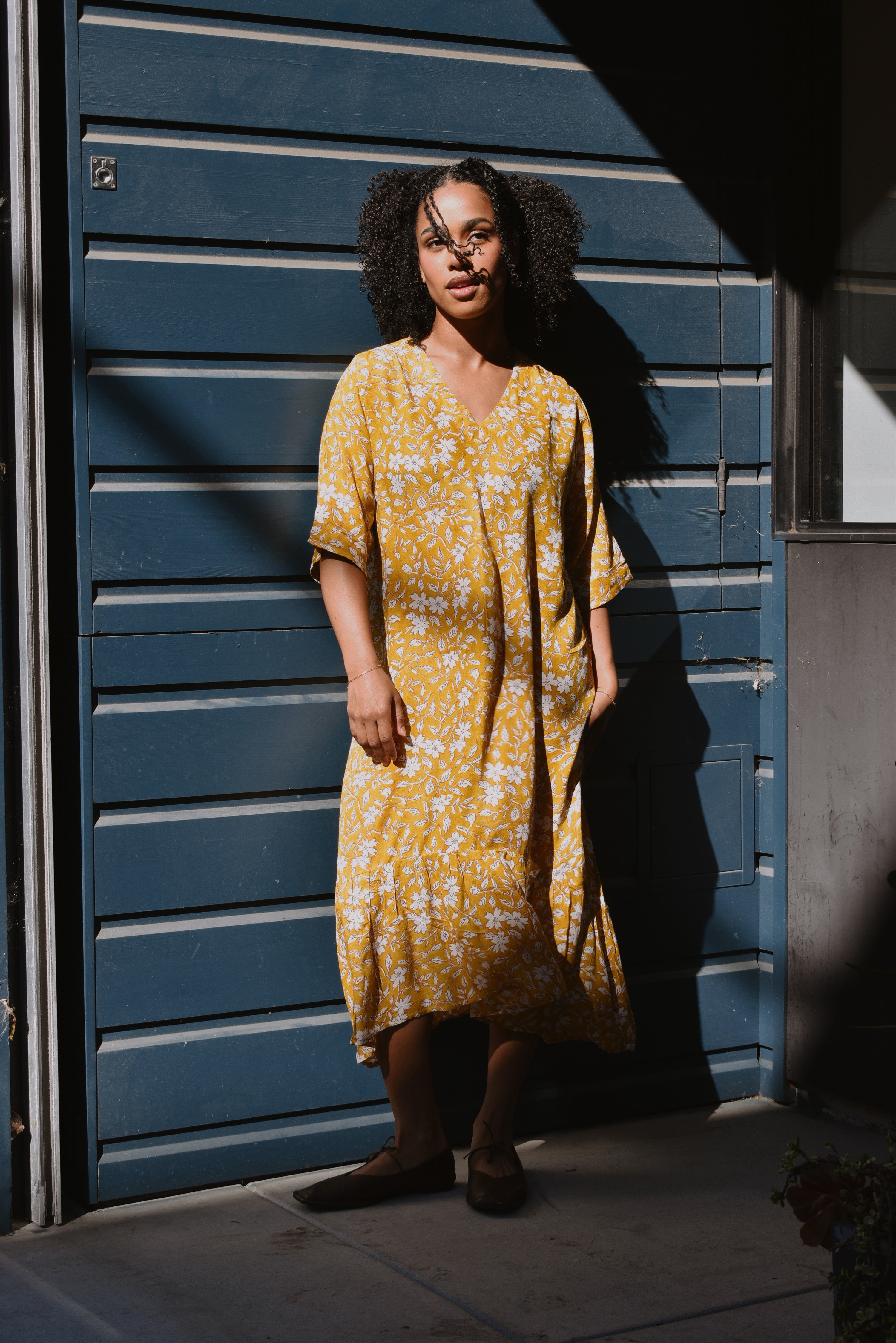 Woman in a yellow floral midi dress standing in front of a blue wall.