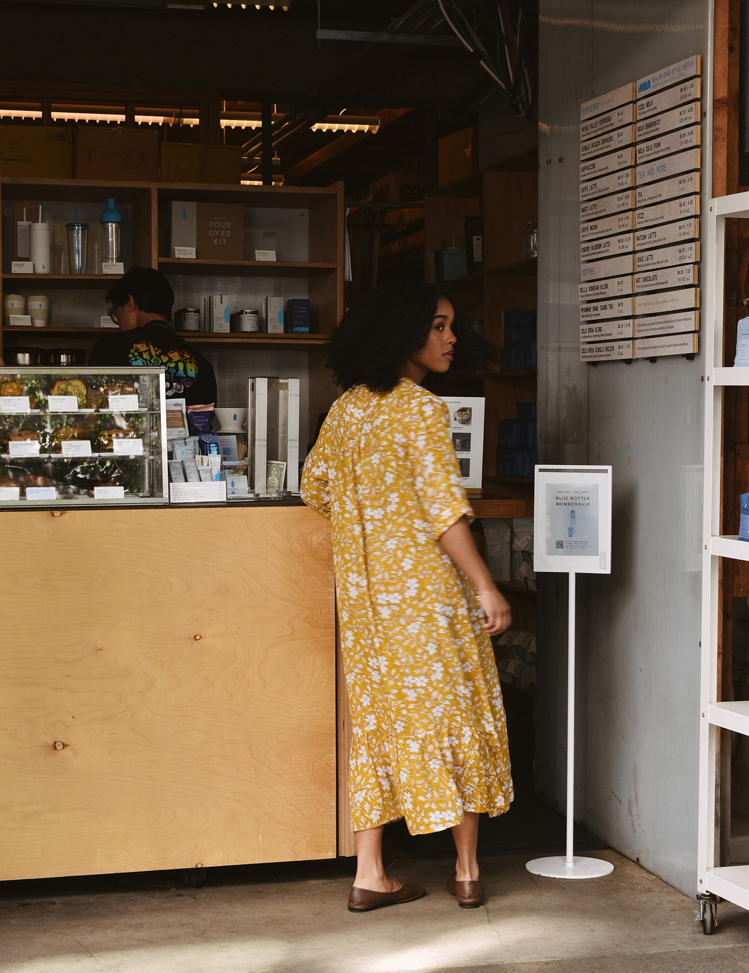 Woman in a yellow floral midi dress standing at a counter in a store.