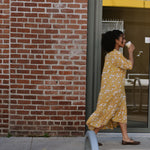 Woman in a yellow floral midi dress walking by a brick wall