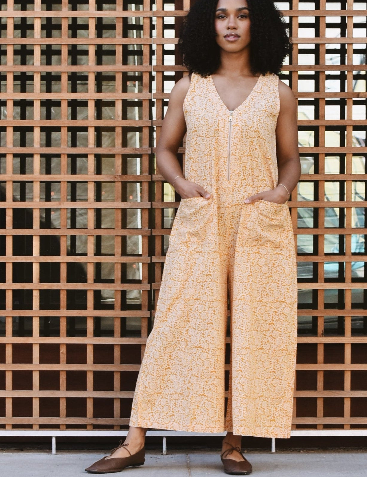 Woman standing against a grid-patterned surface with a building in the background, wearing a yellow floral print jumpsuit