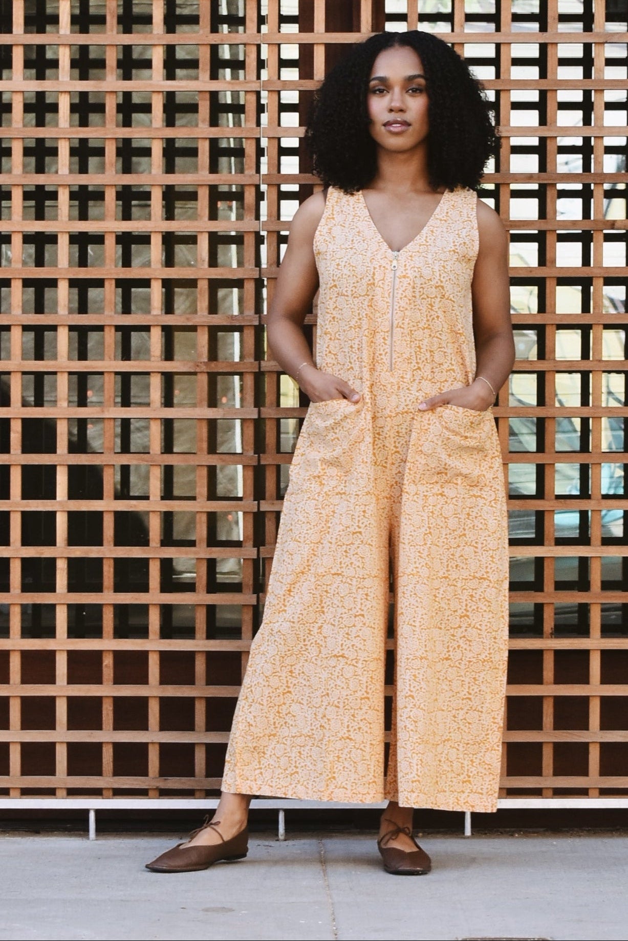 Woman standing against a grid-patterned surface with a building in the background, wearing a yellow floral print jumpsuit