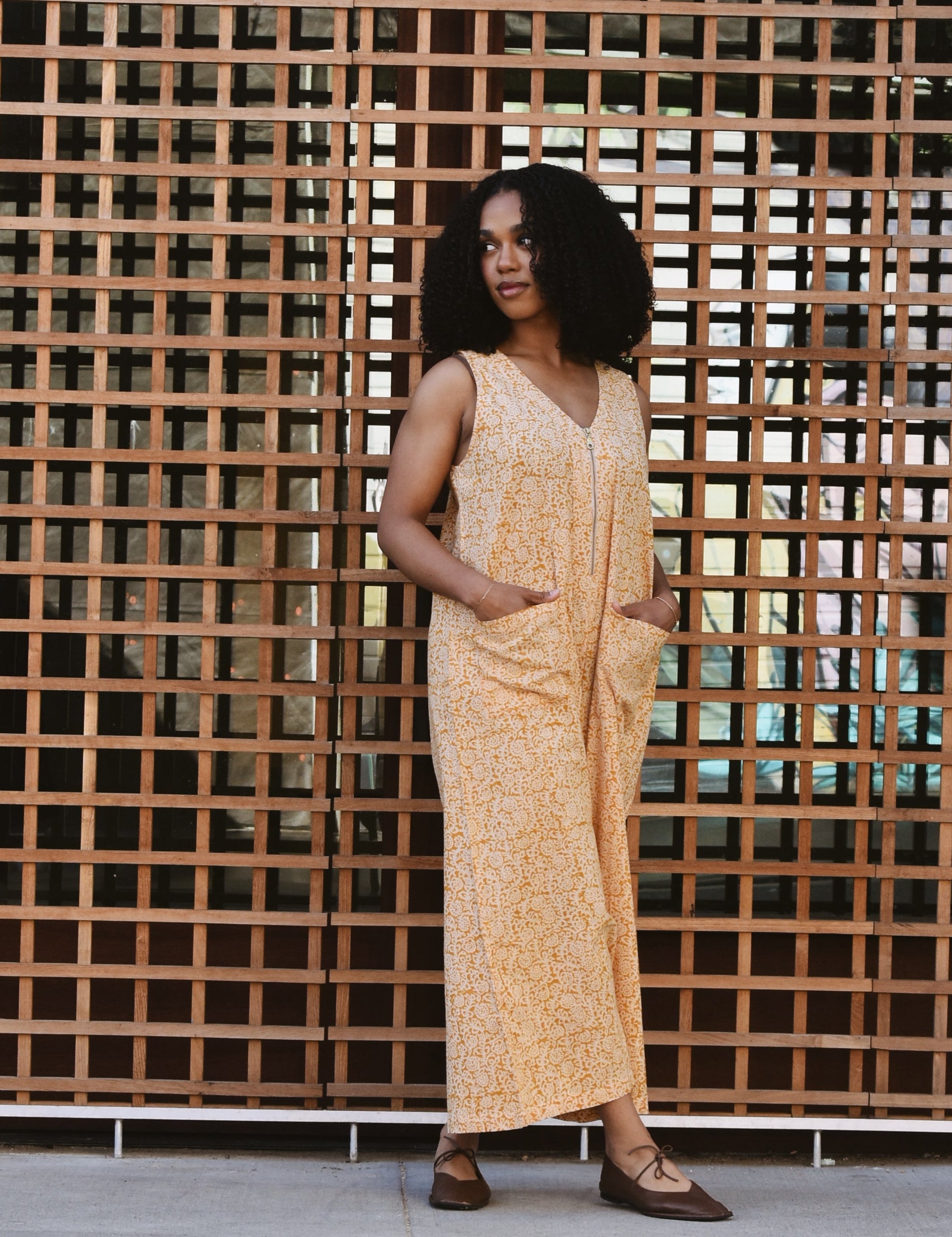 Woman in a yellow floral print jumpsuit standing in front of a grid patterned wall.
