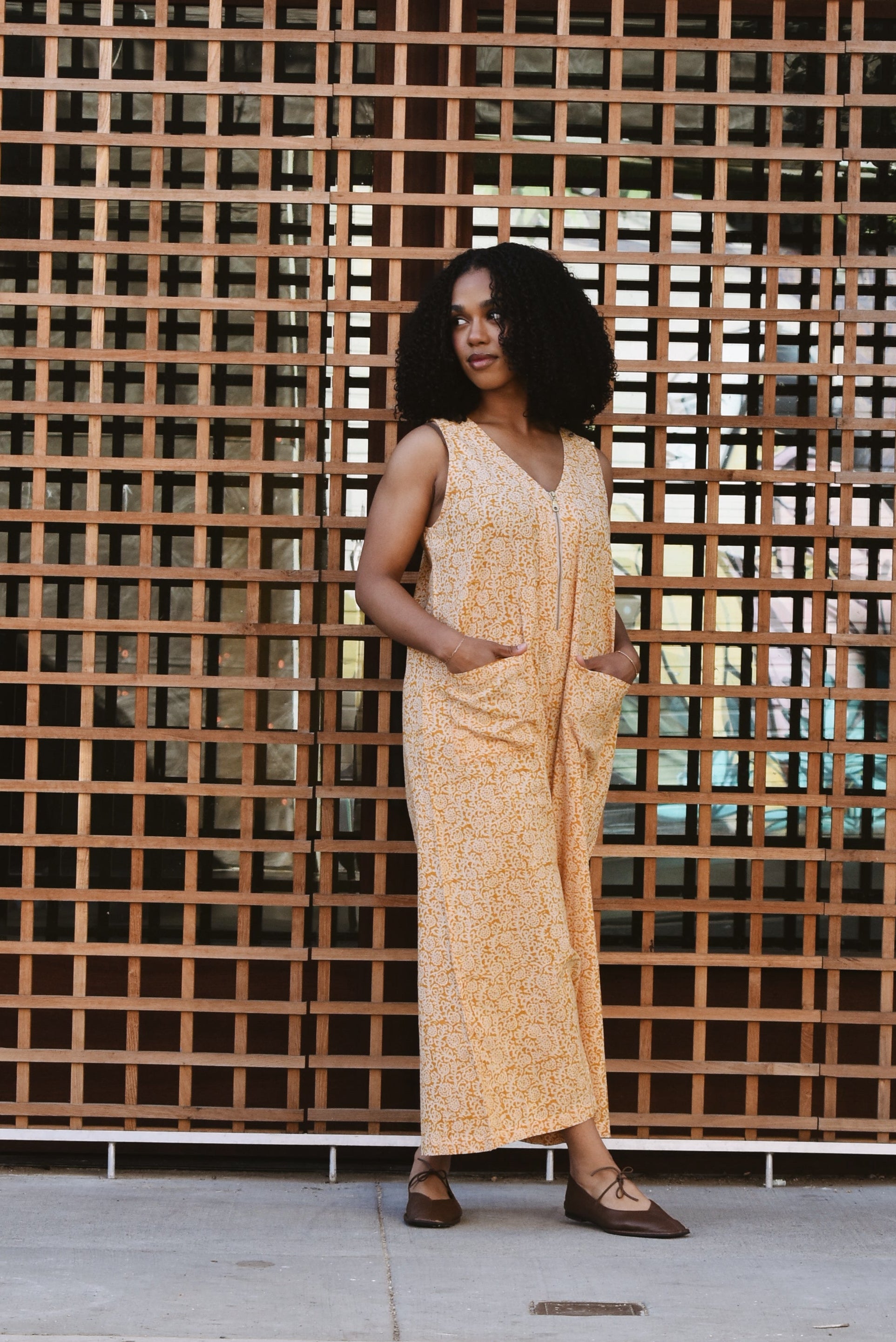 Woman in a yellow floral print jumpsuit standing in front of a grid patterned wall.