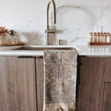 Kitchen sink area with a grayish brown floral print towel hanging on the cabinet, marble backsplash, and wooden cabinets.