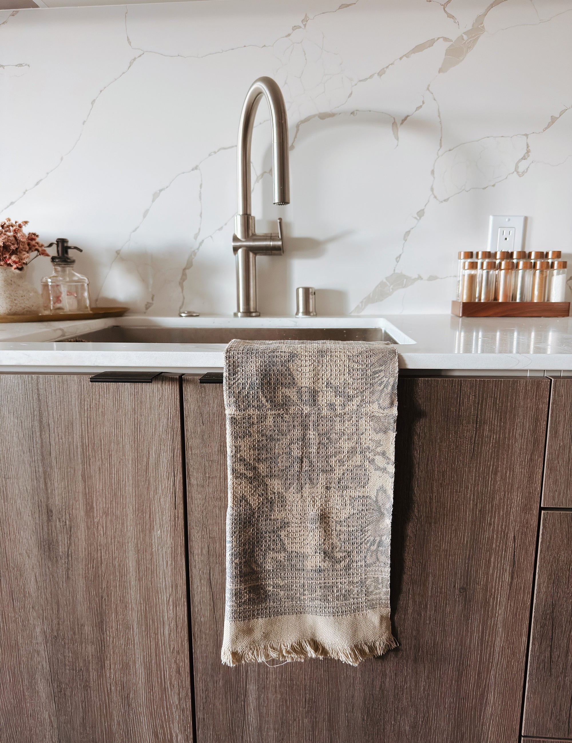 Kitchen sink area with a grayish brown floral print towel hanging on the cabinet, marble backsplash, and wooden cabinets.