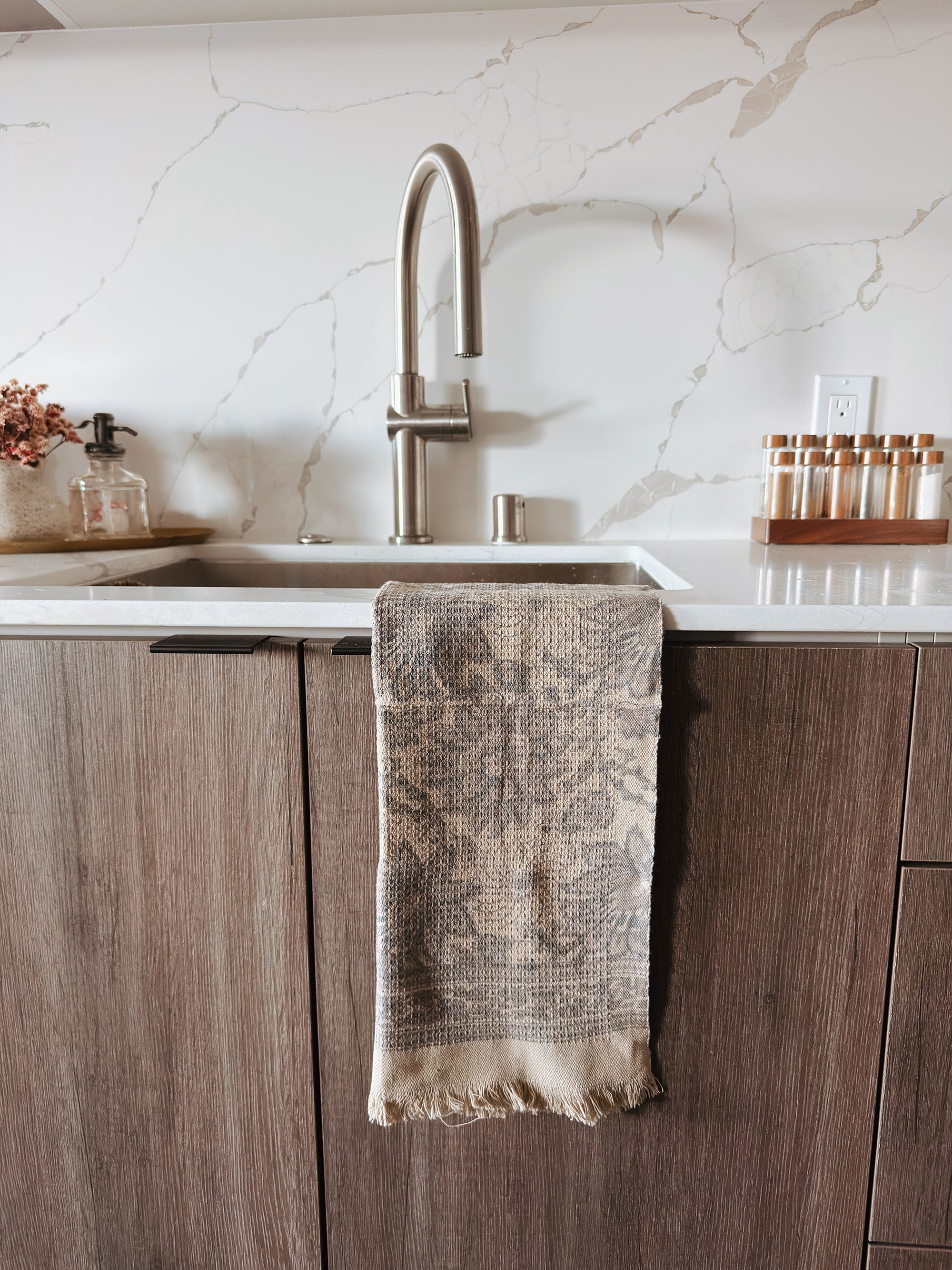 Kitchen sink area with a grayish brown floral print towel hanging on the cabinet, marble backsplash, and wooden cabinets.