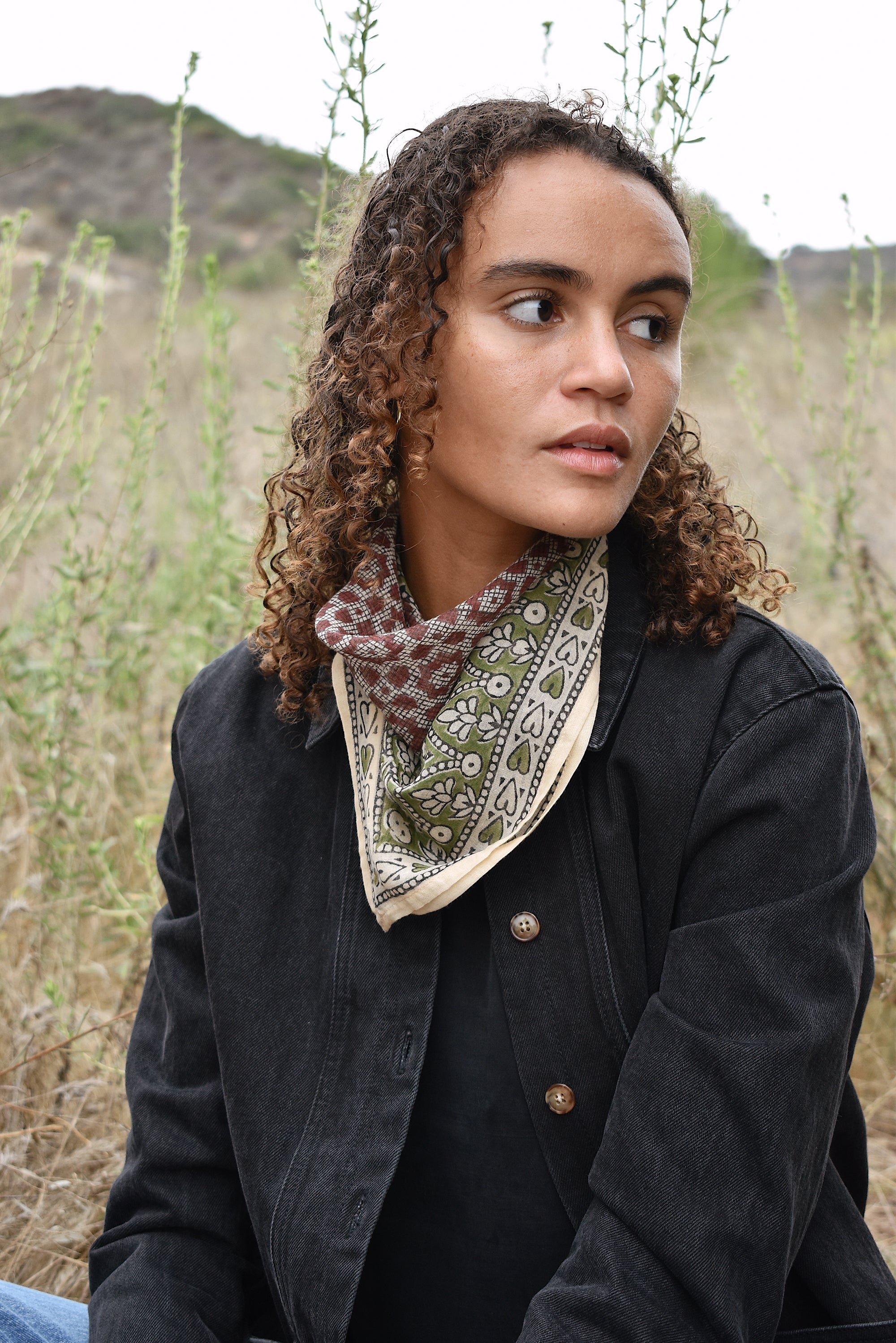 Model wearing Janis bandana, tied in the back around neck, worn with black denim jacket. Model is sitting outside in a field.