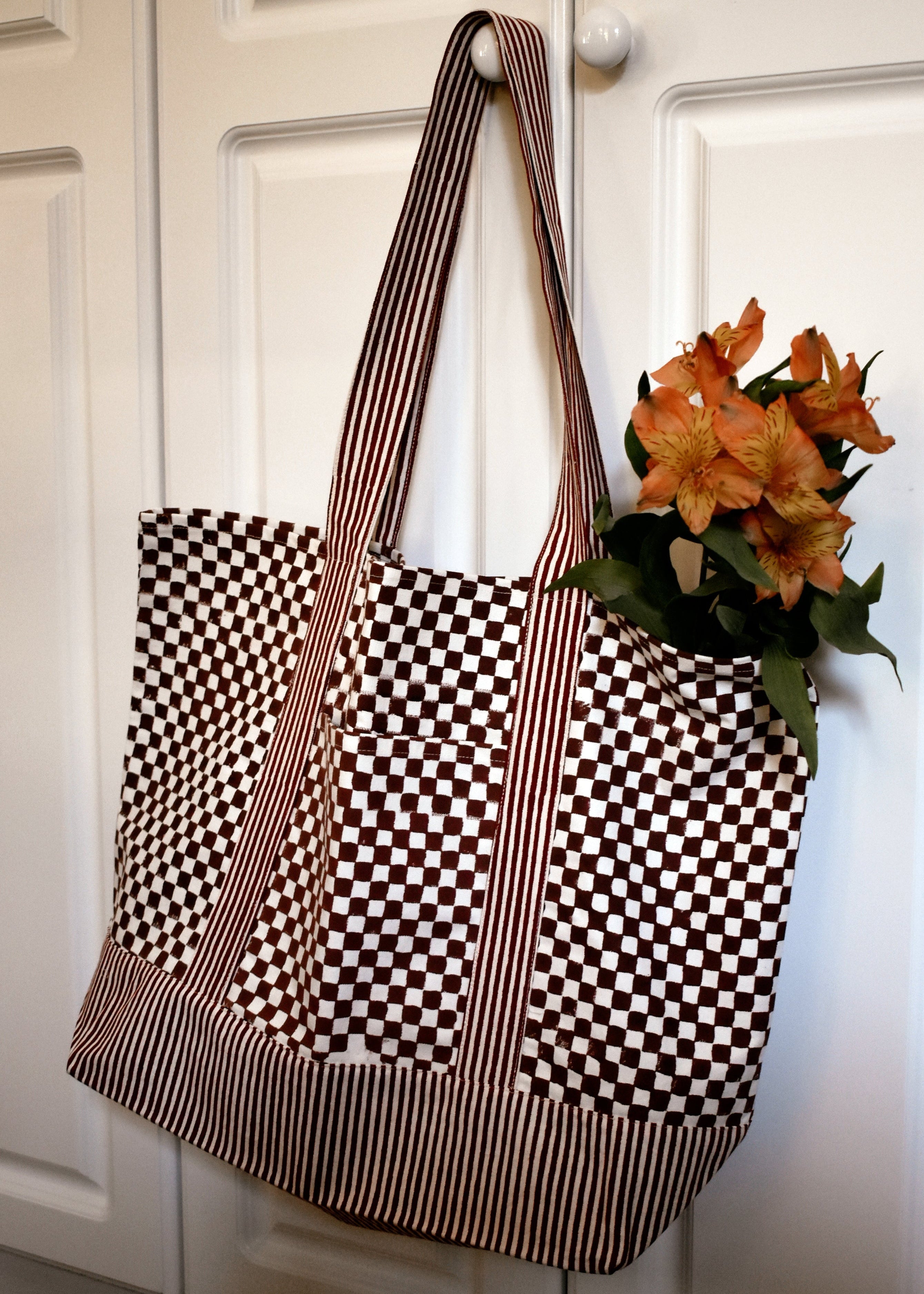Checkered tote bag with striped handles hanging on a white door with flowers hanging out of the bag