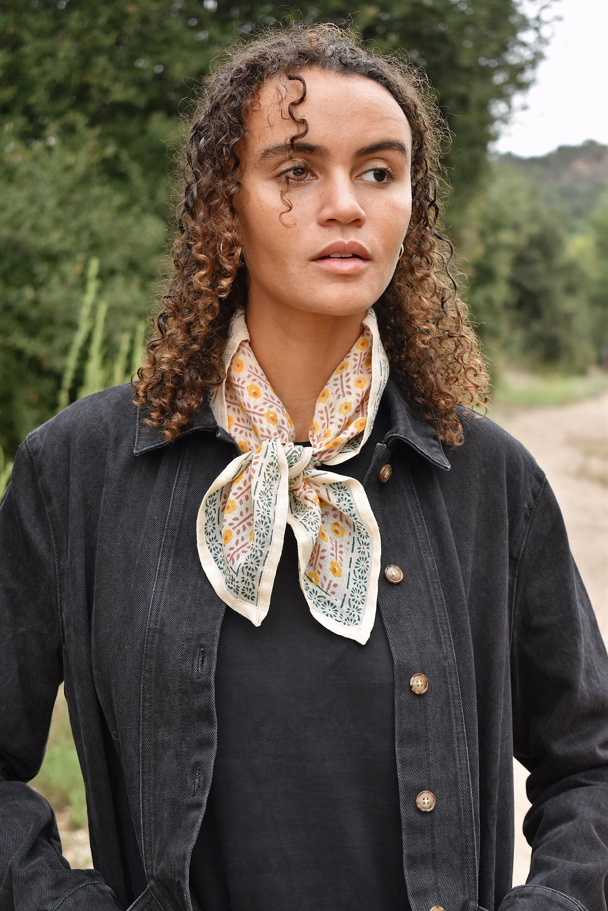 Model wearing Juliet bandana around neck, paired with black denim jacket. Model standing in front of foliage.