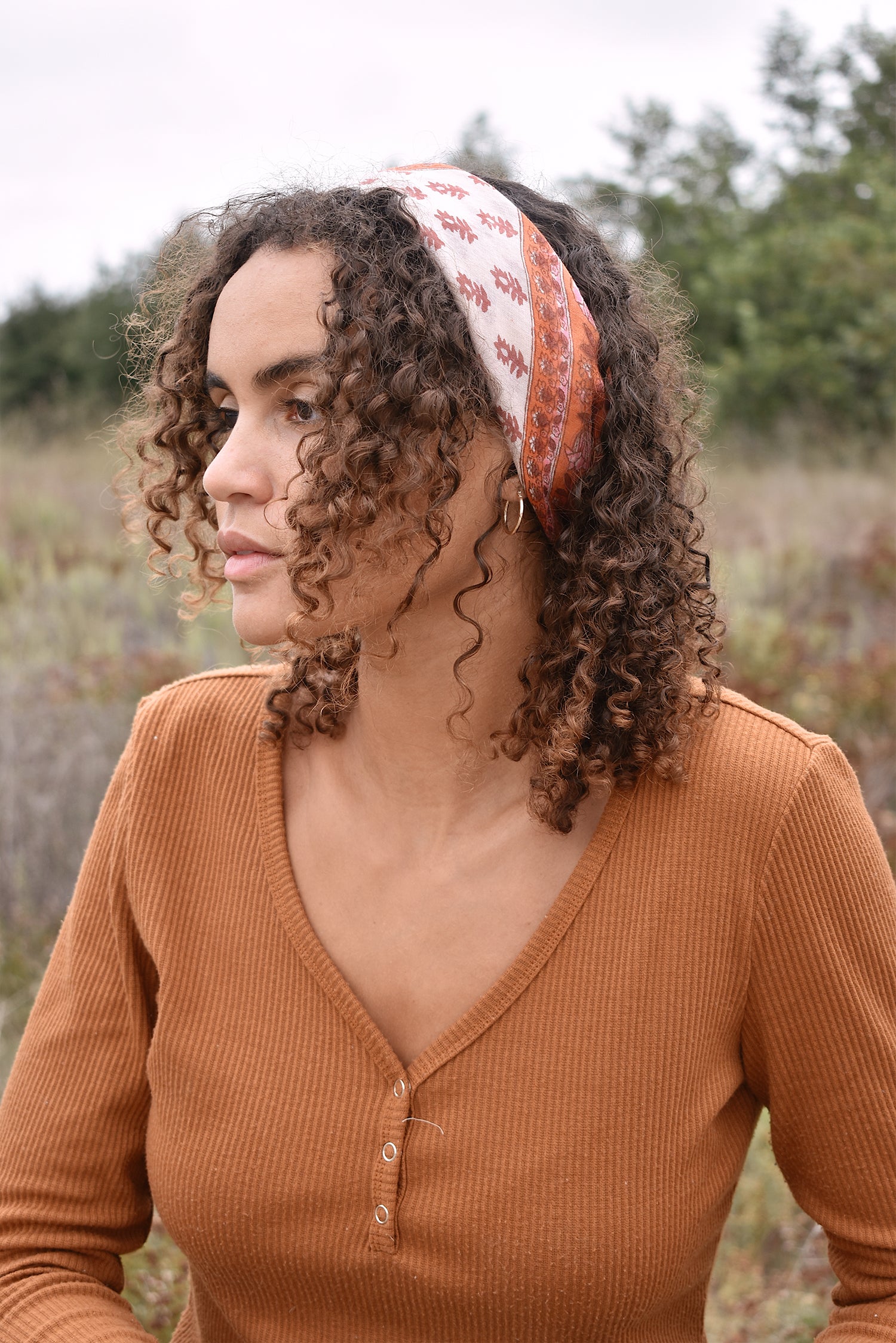 Woman wearing an orange long sleeve top, standing behind a wooden fence with trees in the background, with the block print Rory cotton bandana tied around her hair as a headband.