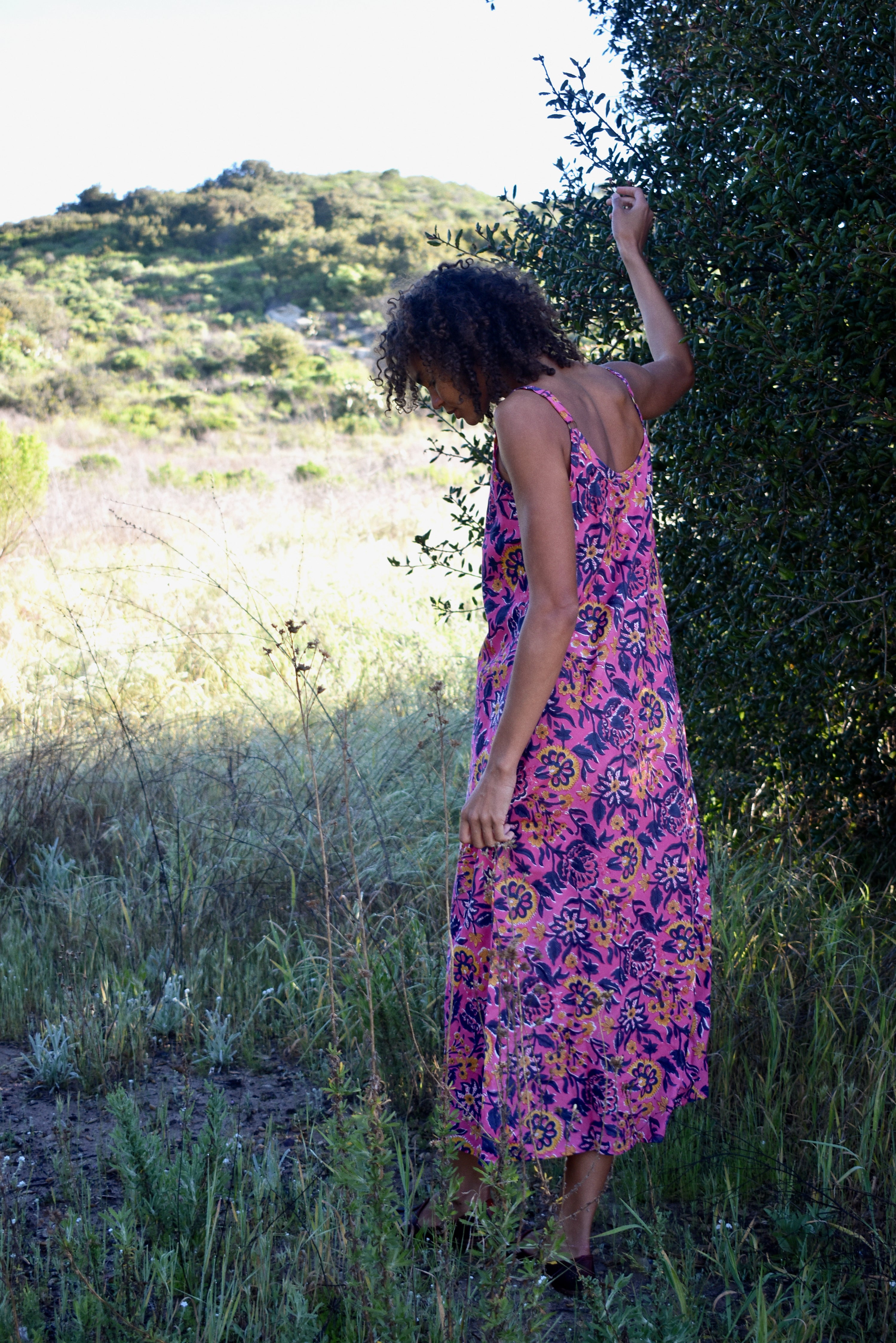 Back shot of woman wearing pink and purple floral printed strappy maxi dress, standing in nature