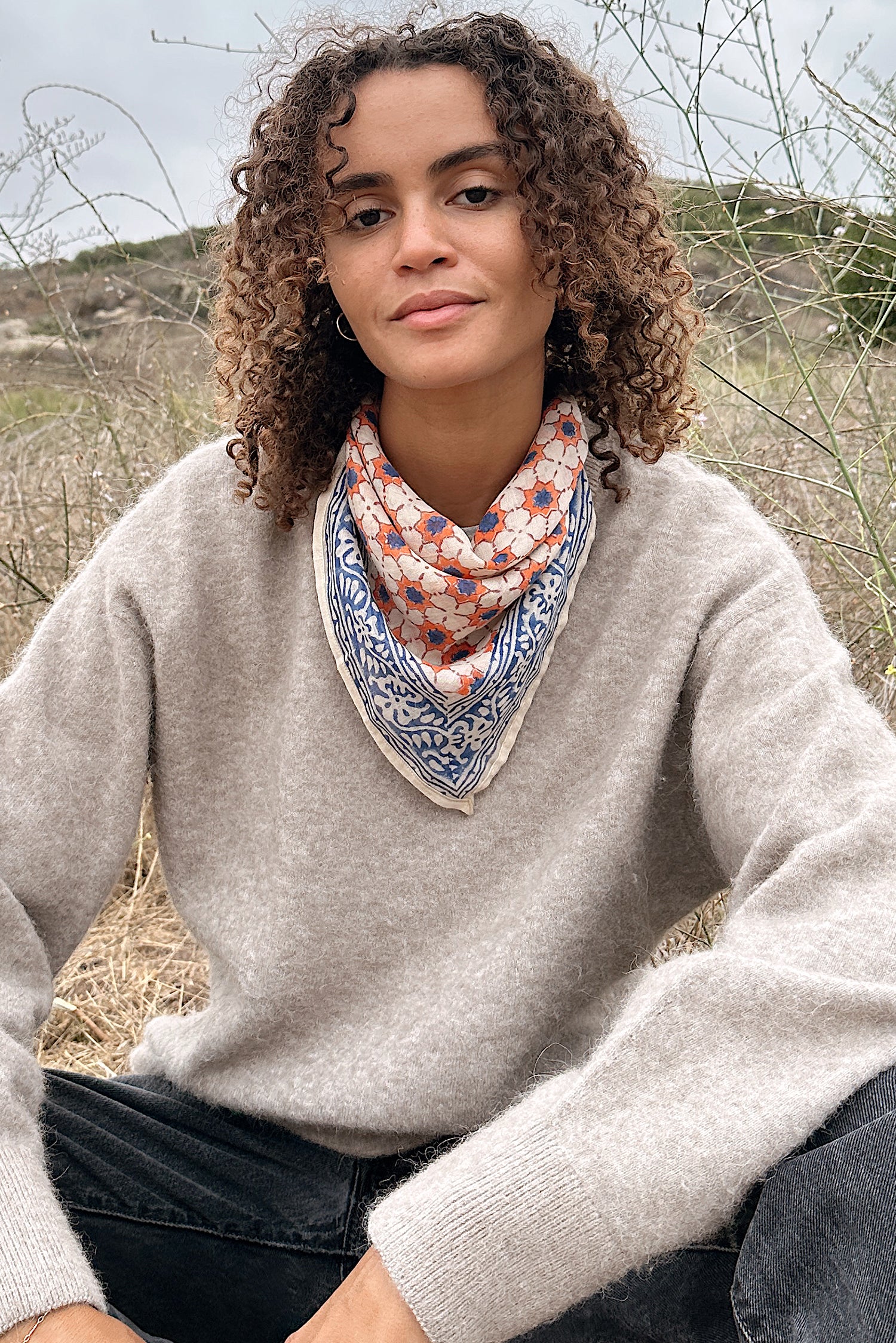 Woman wearing a tan sweater, black jeans, sitting in a field, with the block print Jane cotton bandana tied around her neck.