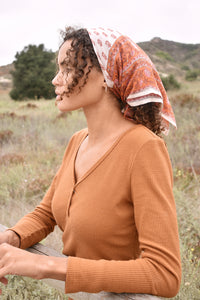 Woman wearing an orange long sleeve top, standing behind a wooden fence with trees in the background, with the block print Rory cotton bandana tied around her hair.