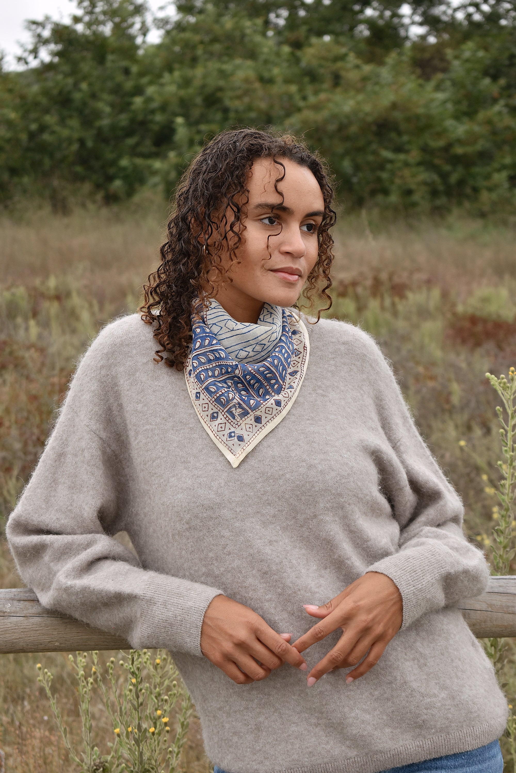Model wearing Helene bandana tied around neck, tied in the back. Paired with tan sweater and blue jeans. Model leaning against a fence, outside near trees.