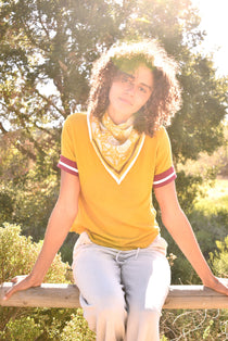 Woman wearing block print Tate bandana around the neck against a yellow tee shirt. Woman sitting on wooden fence with trees in the background.