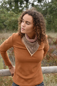 Woman wearing an orange long sleeve top, standing in front of trees, with the block print Theo cotton bandana tied around her neck. Woman is leaning forward.