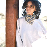 Woman wearing block print Frontier bandana tied around neck against a grey sweatshirt. Woman leaning against wooden pillar.