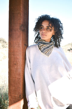 Woman wearing block print Frontier bandana tied around neck against a grey sweatshirt. Woman leaning against wooden pillar.