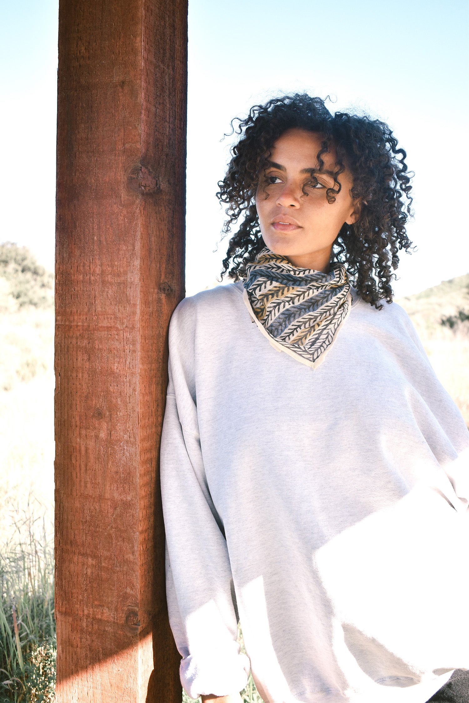 Woman wearing block print Frontier bandana tied around neck against a grey sweatshirt. Woman leaning against wooden pillar.